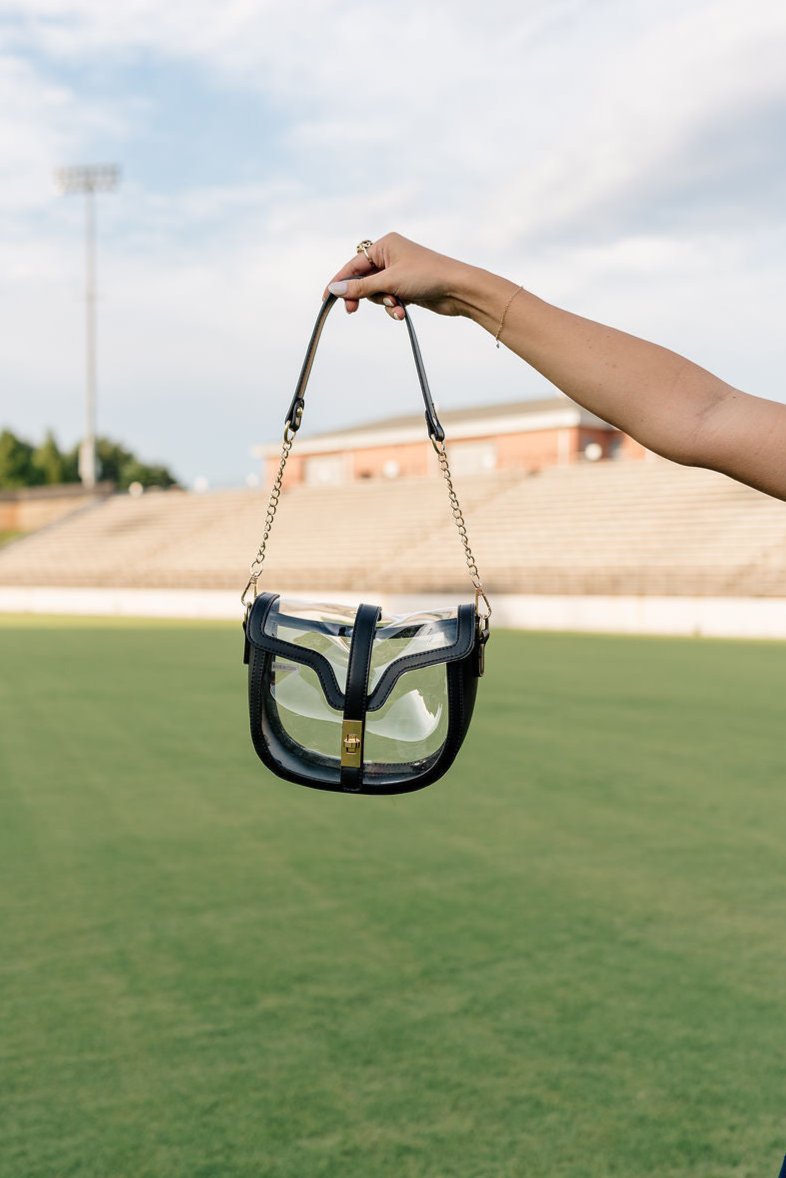 Image shows model's hand holding the Margaret Clear & Black Purse in front of grass field.