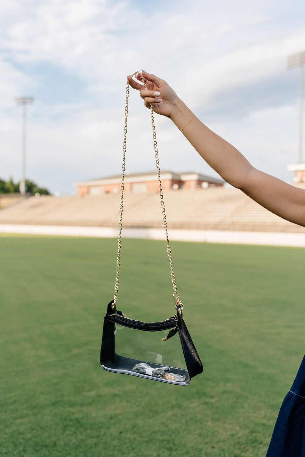 Model's hand is holding the Rebecca Clear Crossbody Purse in Black in front of a grass field background.