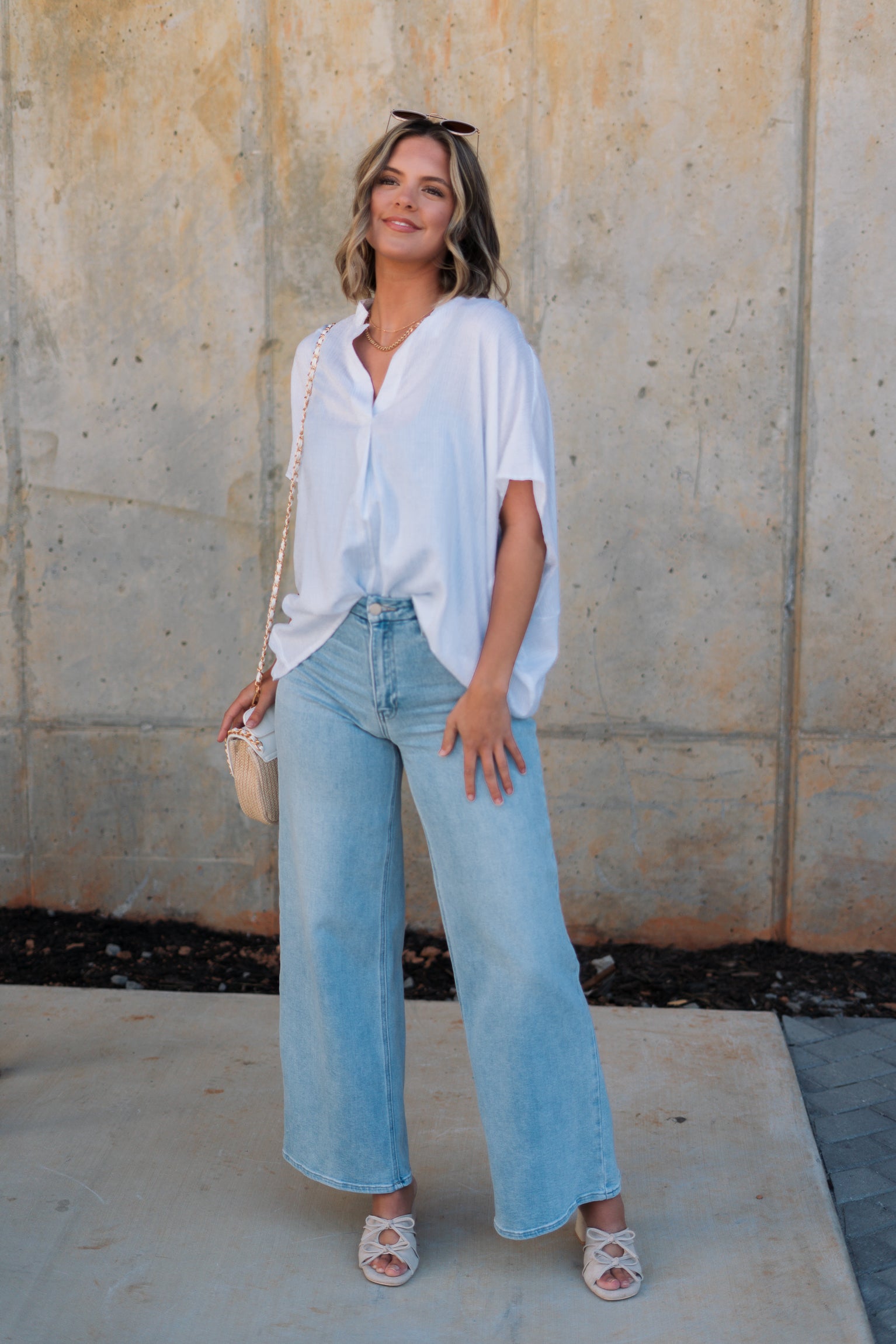 Full body front view of female model wearing the Thea White Lightweight Top that has lightweight white fabric with subtle monochrome stripes, oversized fit, v neckline, and short batwing sleeves. Shirt is front tucked into jeans.