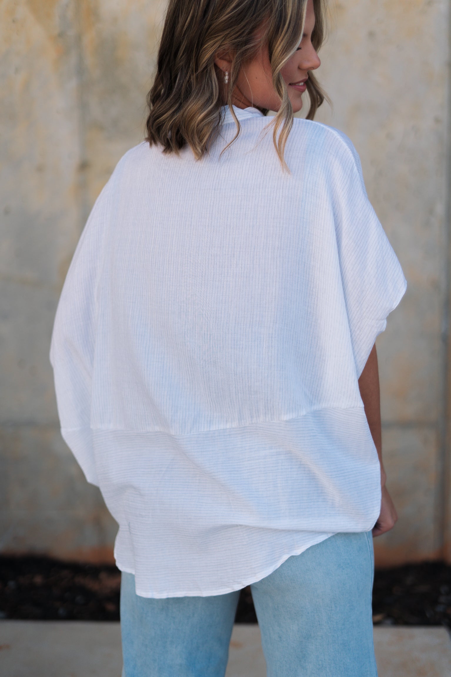 Back view of female model wearing the Thea White Lightweight Top that has lightweight white fabric with subtle monochrome stripes, oversized fit, v neckline, and short batwing sleeves.