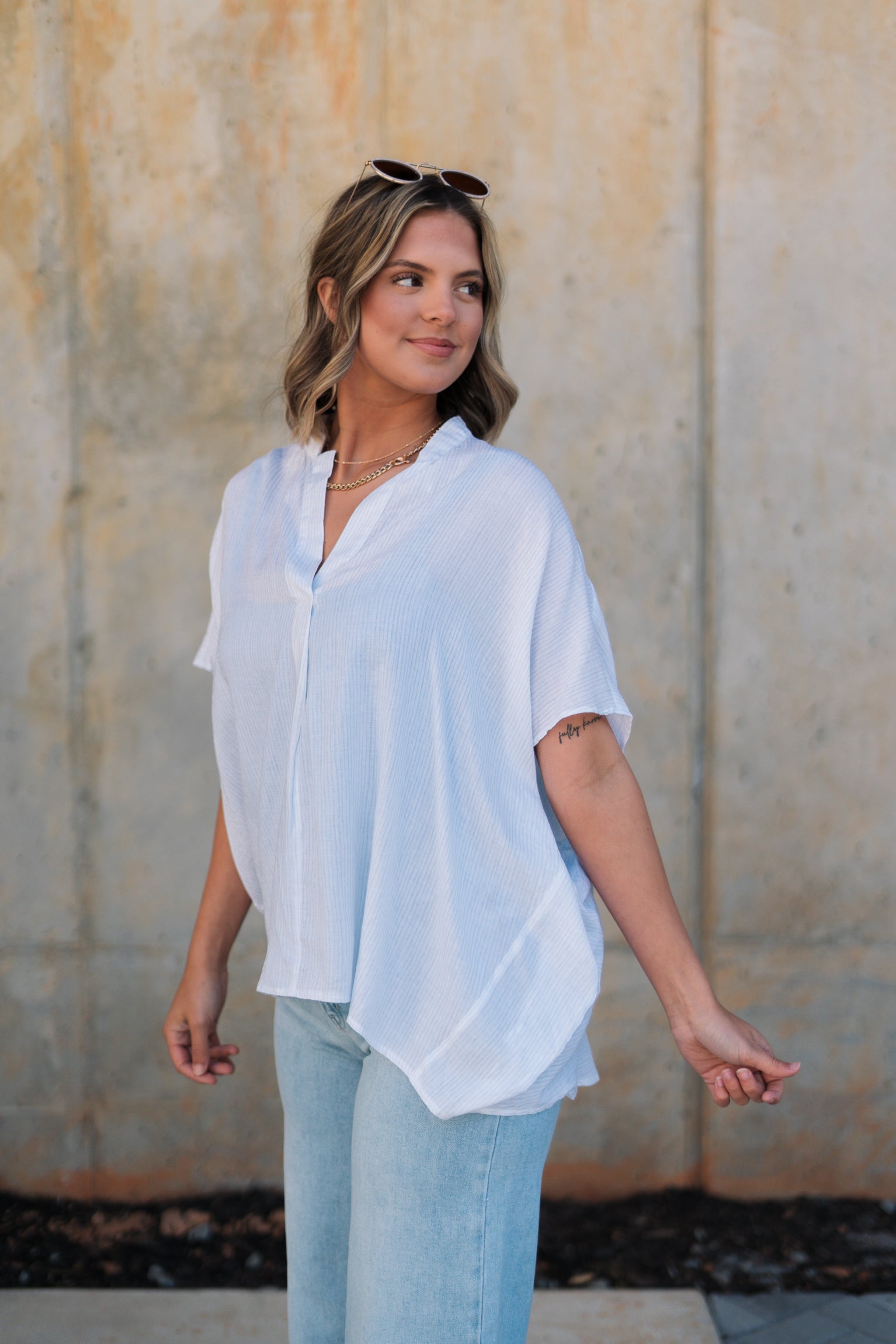 Frontal side view of female model wearing the Thea White Lightweight Top that has lightweight white fabric with subtle monochrome stripes, oversized fit, v neckline, and short batwing sleeves.