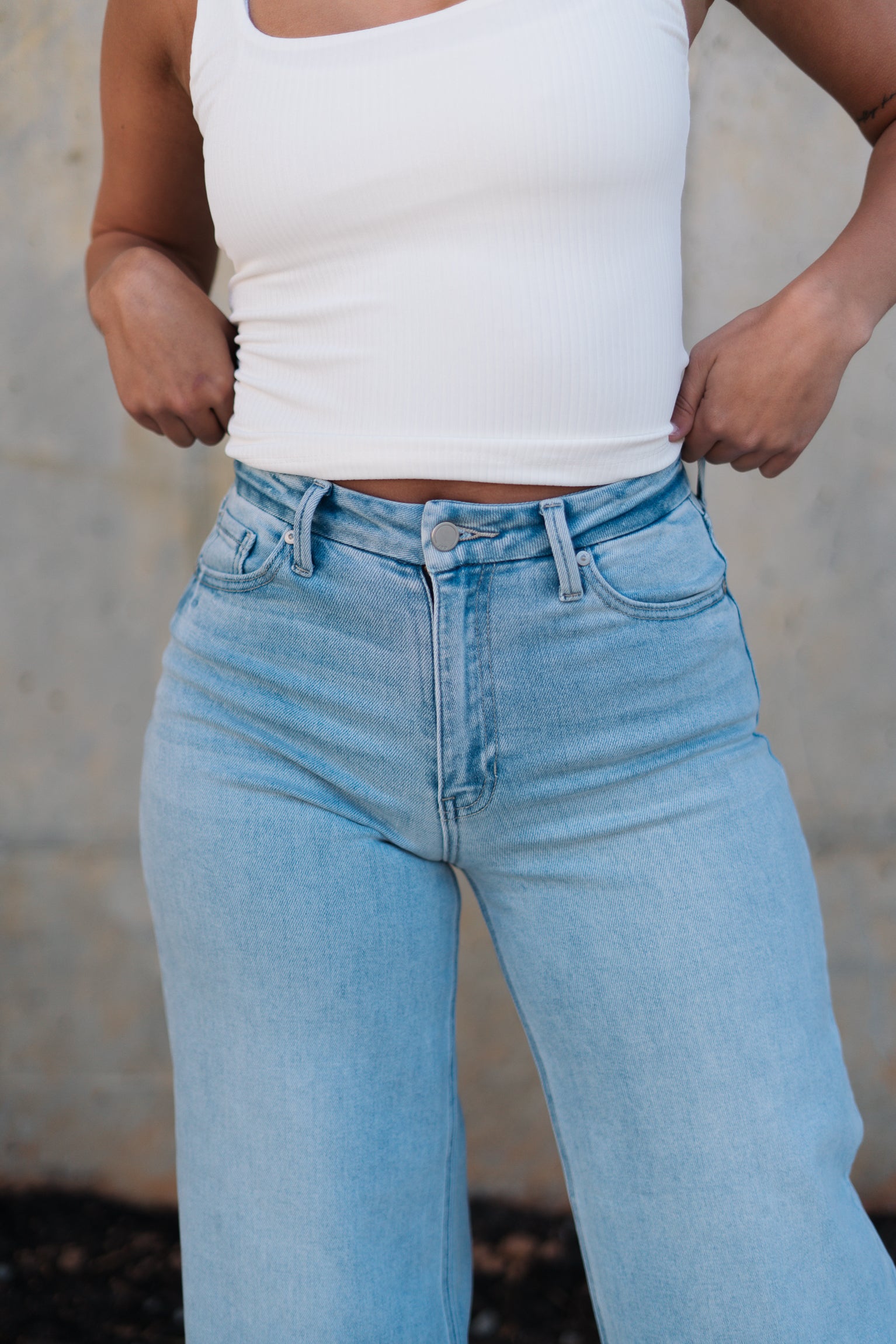 Upper body front view of female model wearing the Kristen Light Wash Wide Leg Jeans that features wide cropped legs, light wash denim, belt loops, pockets, and a zipper/button fly. Worn with white tank top