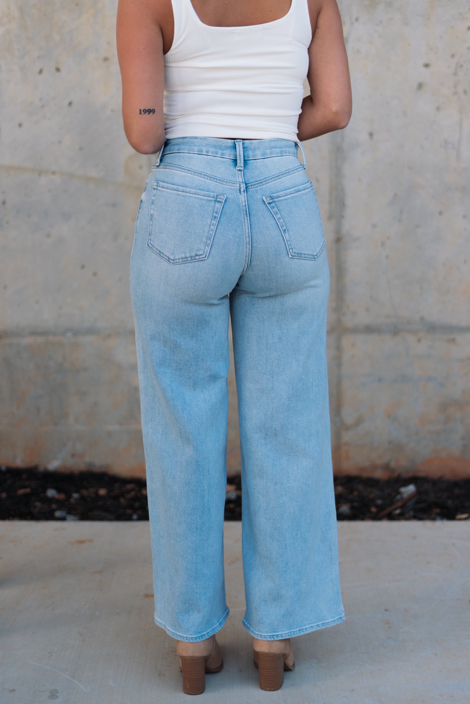 Back view of female model wearing the Kristen Light Wash Wide Leg Jeans that features wide cropped legs, light wash denim, belt loops, pockets, and a zipper/button fly. Worn with white tank top