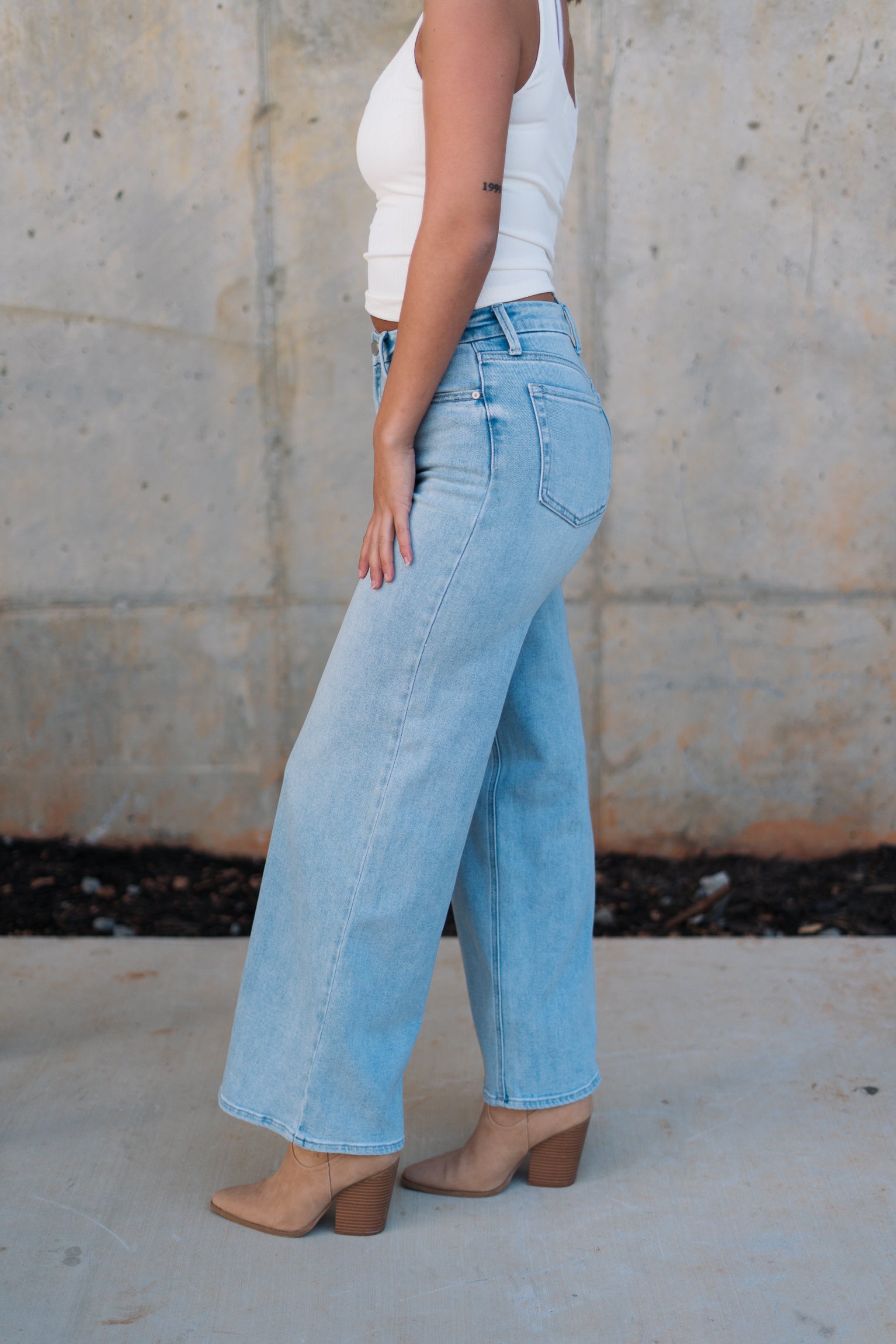 Side view of female model wearing the Kristen Light Wash Wide Leg Jeans that features wide cropped legs, light wash denim, belt loops, pockets, and a zipper/button fly. Worn with white tank top