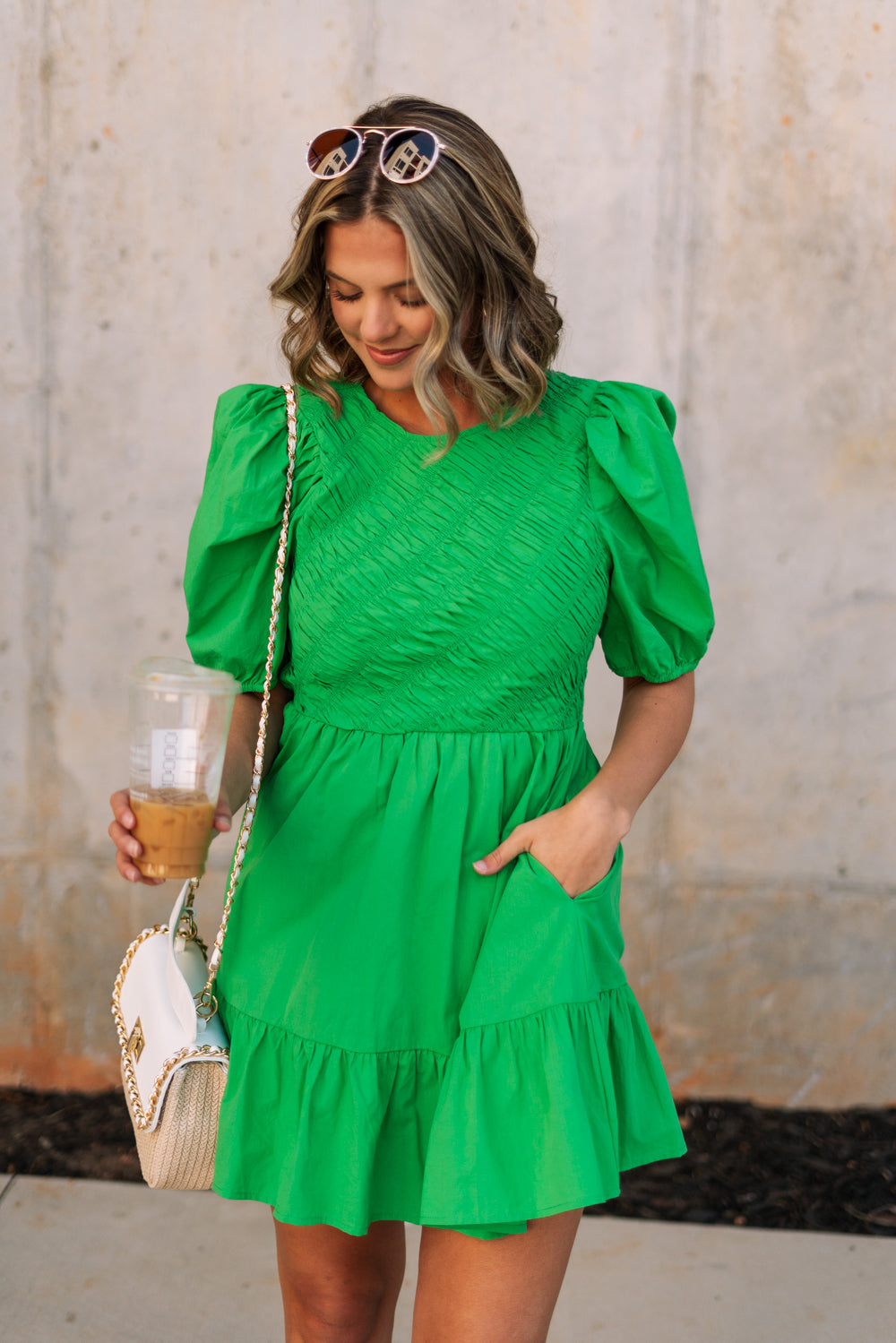 front view of female model wearing the Lea Green Puff Sleeve Mini Dress that has kelly green fabric, a smocked upper with high neck, short puff sleeves, and mini length. Model is holding coffee and has purse on shoulder.