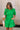 front view of female model wearing the Lea Green Puff Sleeve Mini Dress that has kelly green fabric, a smocked upper with high neck, short puff sleeves, and mini length. Model is holding coffee and has purse on shoulder.