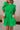 Close-up front view of female model wearing the Lea Green Puff Sleeve Mini Dress that has kelly green fabric, a smocked upper with high neck, short puff sleeves, and mini length. Model is holding coffee and has purse on shoulder.