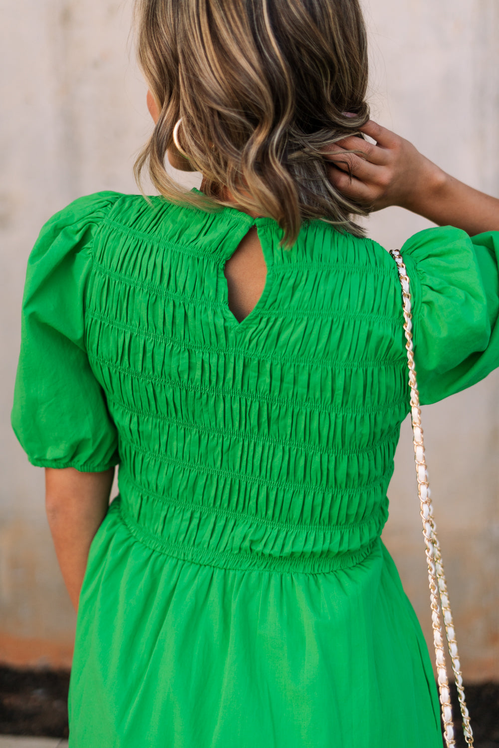 Upper body back view of female model wearing the Lea Green Puff Sleeve Mini Dress that has kelly green fabric, a smocked upper with high neck, short puff sleeves, and mini length. Model is holding coffee and has purse on shoulder.