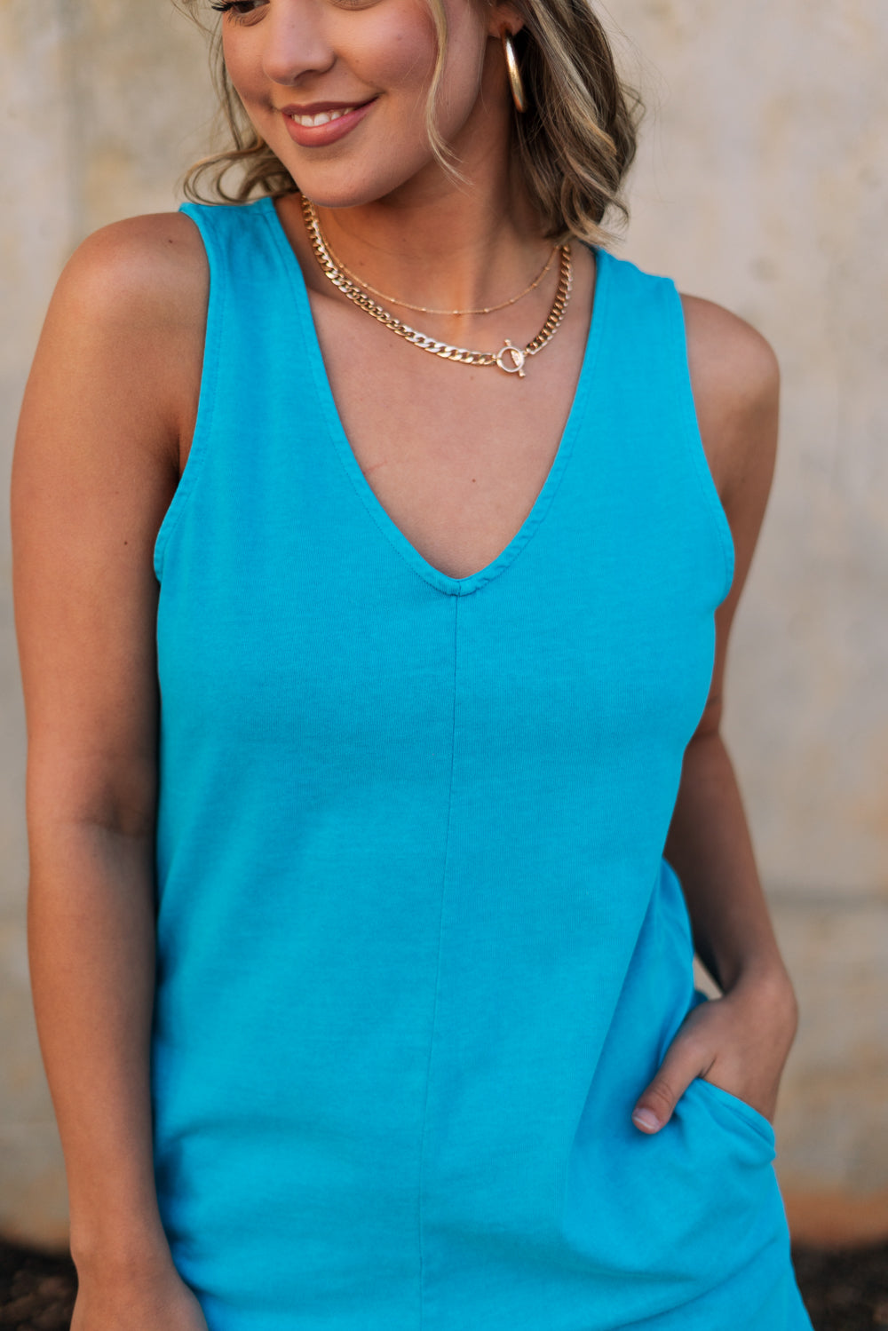 Upper body front view of female model wearing the Chelsea Blue Sleeveless Romper that has bright blue fabric, a v neck, side pockets, and thick straps. Model has hands in pockets.