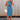 Full body front view of female model wearing the Paula Blue Ribbed Midi Dress that has ribbed medium blue fabric, a round neck, sleeveless, and midi length hem. Model has beige purse on shoulder.