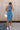 Full body front view of female model wearing the Paula Blue Ribbed Midi Dress that has ribbed medium blue fabric, a round neck, sleeveless, and midi length hem. Model has beige purse on shoulder.