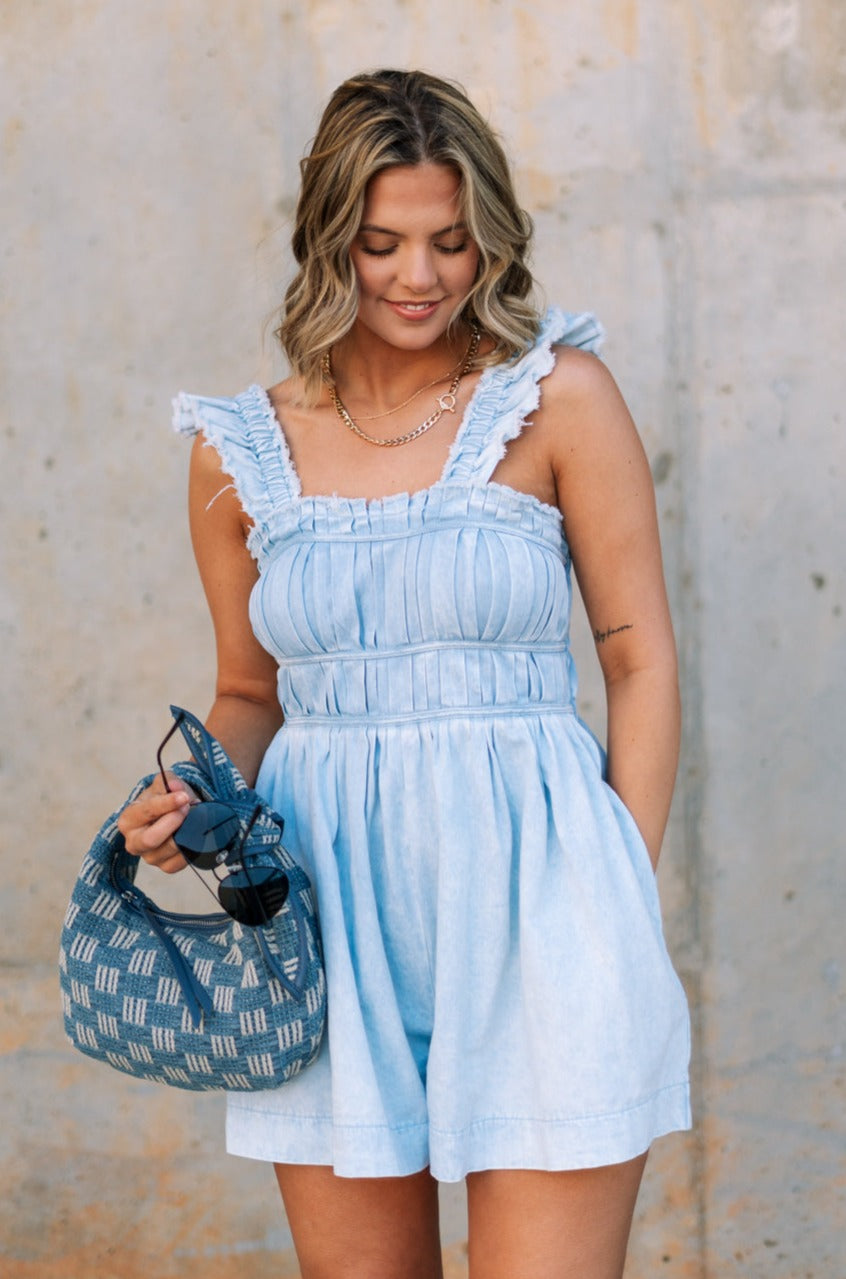 front view of female model wearing the Taylor Light Wash Denim Romper that has light wash denim fabric, straps with frayed hems, elastic waist, neckline pleating, and side pockets.