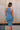 Back view of female model wearing the Paula Blue Ribbed Midi Dress that has ribbed medium blue fabric, a round neck, sleeveless, and midi length hem. Model has beige purse on shoulder.