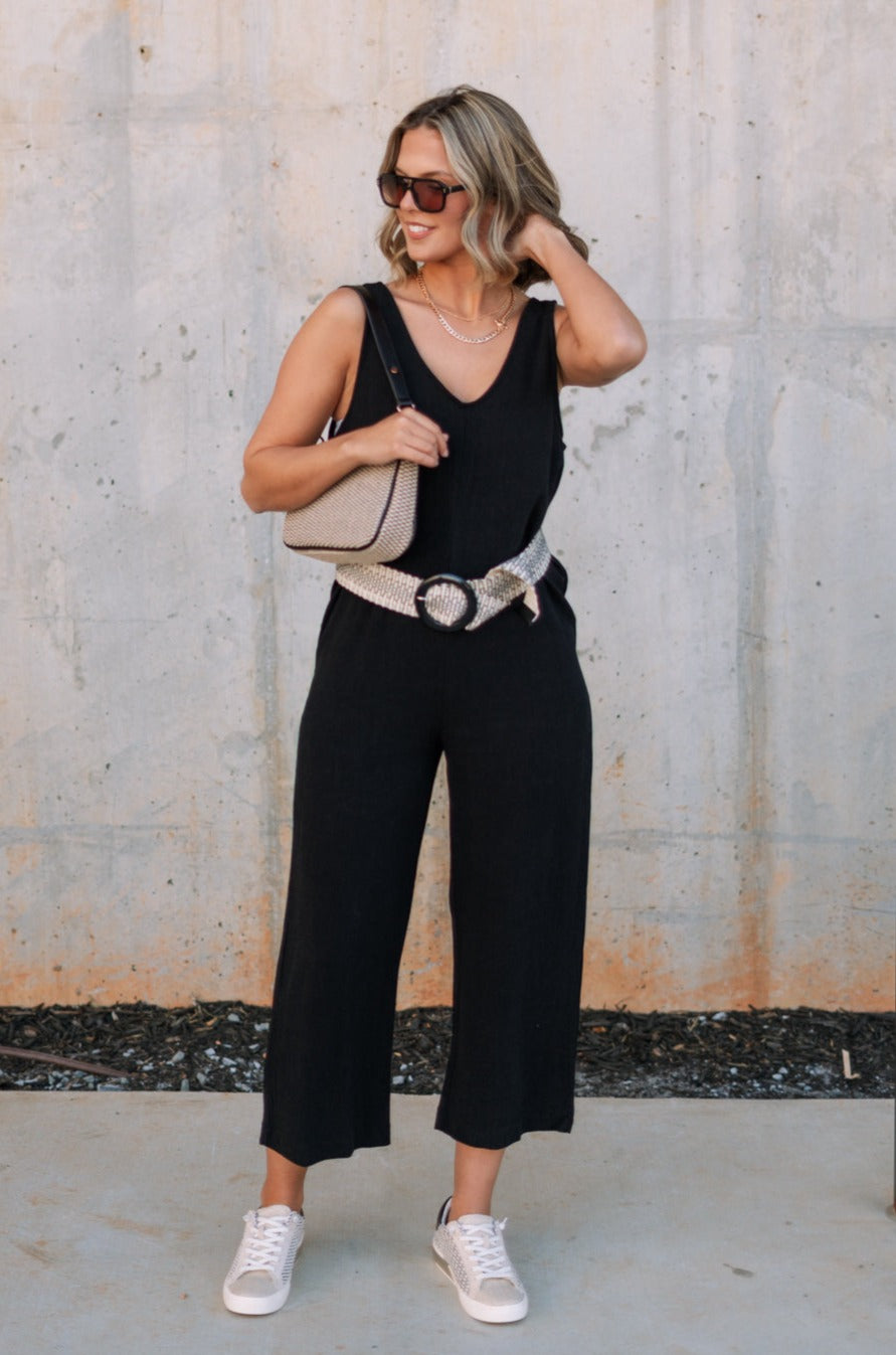 Full body front view of female model wearing the Kinsley Black Sleeveless Jumpsuit that has black lightweight fabric, thick straps, wide legs, and a v-neck. Styled with a belt and a purse on model's shoulder.