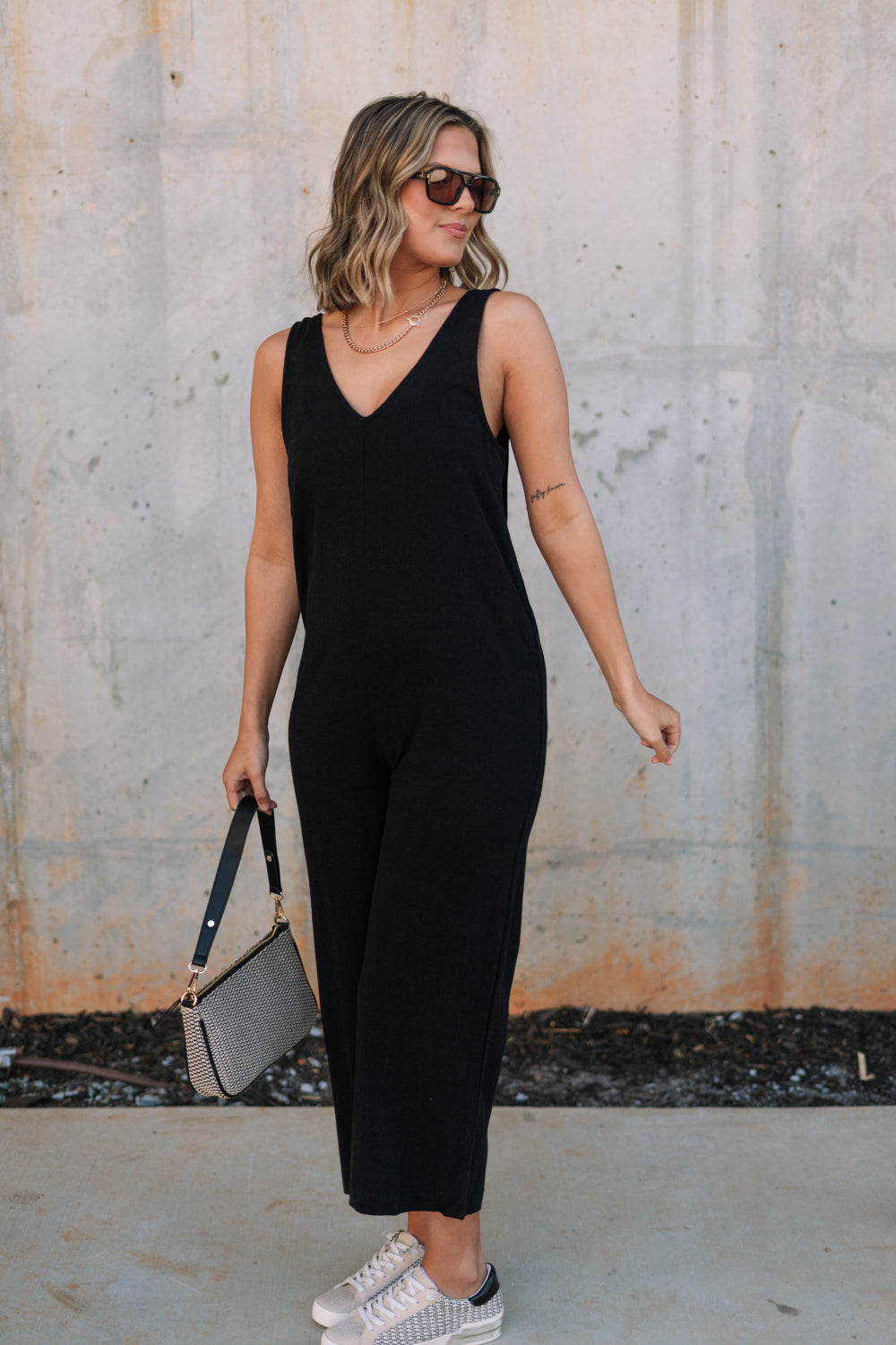 Full body side view of female model wearing the Kinsley Black Sleeveless Jumpsuit that has black lightweight fabric, thick straps, wide legs, and a v-neck. 