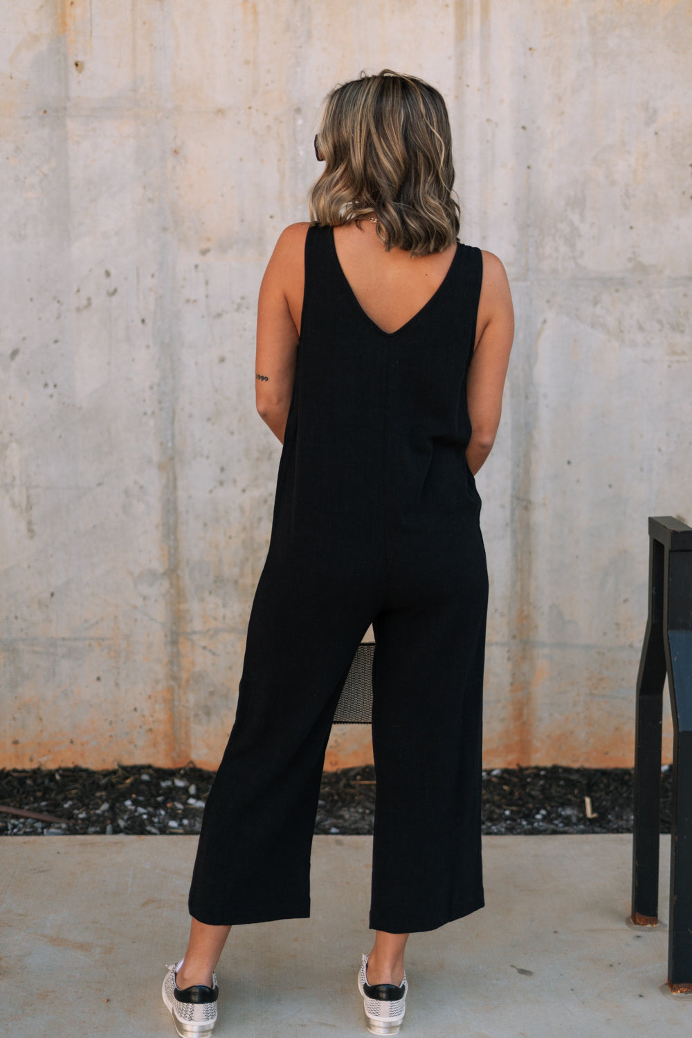 Full body back view of female model wearing the Kinsley Black Sleeveless Jumpsuit that has black lightweight fabric, thick straps, wide legs, and a v-neck. 
