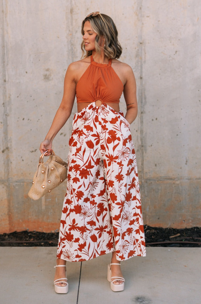 Full body front view of female model wearing the Cali Rust & Cream Cutout Floral Jumpsuit that has a rust upper with a halter neckline, midriff cutouts with a ring in center, and white wide leg pants with rust floral print.