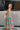 Full body side view of female model wearing the Leilani Printed Sleeveless Maxi Dress that has a multi-colored green, red, pink, purple, and yellow print, spaghetti straps, a smocked upper, and a tiered skirt.