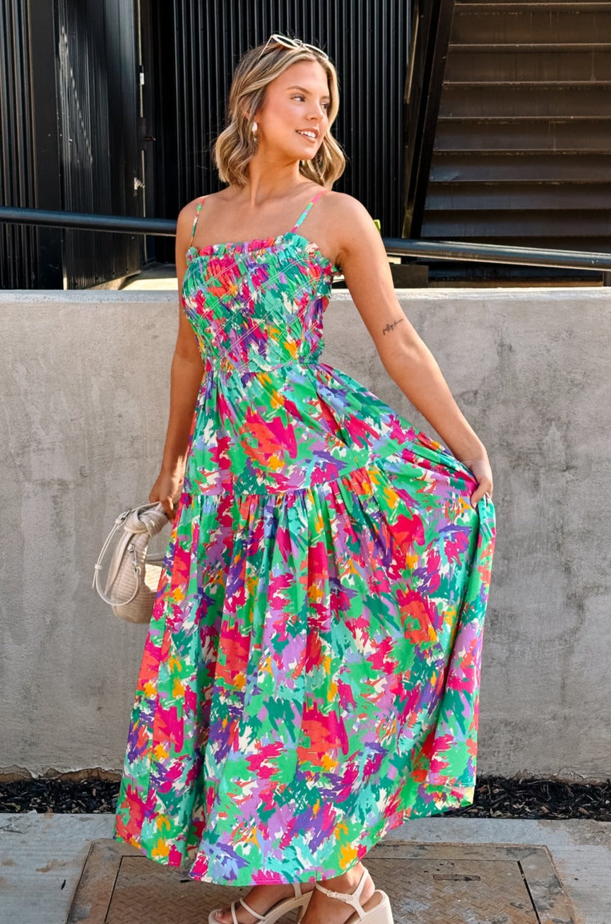 Full body front view of female model wearing the Leilani Printed Sleeveless Maxi Dress that has a multi-colored green, red, pink, purple, and yellow print, spaghetti straps, a smocked upper, and a tiered skirt.