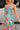 Close-up front/side view of female model wearing the Leilani Printed Sleeveless Maxi Dress that has a multi-colored green, red, pink, purple, and yellow print, spaghetti straps, a smocked upper, and a tiered skirt.