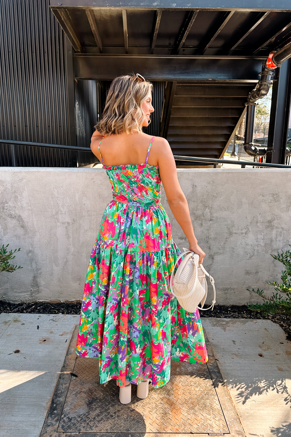 Full body back view of female model wearing the Leilani Printed Sleeveless Maxi Dress that has a multi-colored green, red, pink, purple, and yellow print, spaghetti straps, a smocked upper, and a tiered skirt.