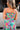 Upper body back view of female model wearing the Leilani Printed Sleeveless Maxi Dress that has a multi-colored green, red, pink, purple, and yellow print, spaghetti straps, a smocked upper, and a tiered skirt.