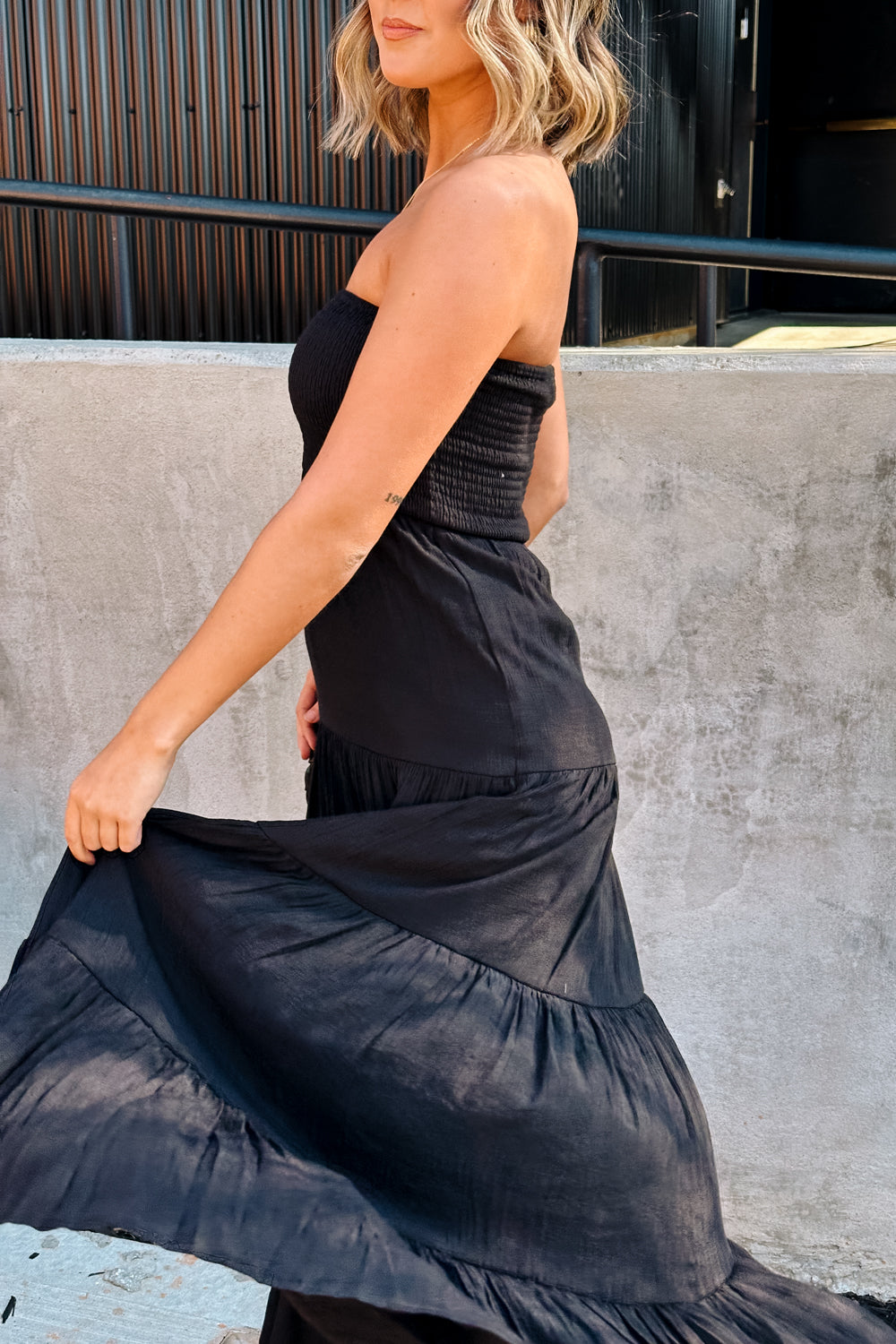 Side view of female model wearing the Amaya Black Strapless Maxi Dress that has a smocked upper and a tiered skirt. Model is holding skirt
