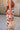 Close-up side view of female model wearing the Cali Rust & Cream Cutout Floral Jumpsuit that has a rust upper with a halter neckline, midriff cutouts with a ring in center, and white wide leg pants with rust floral print.