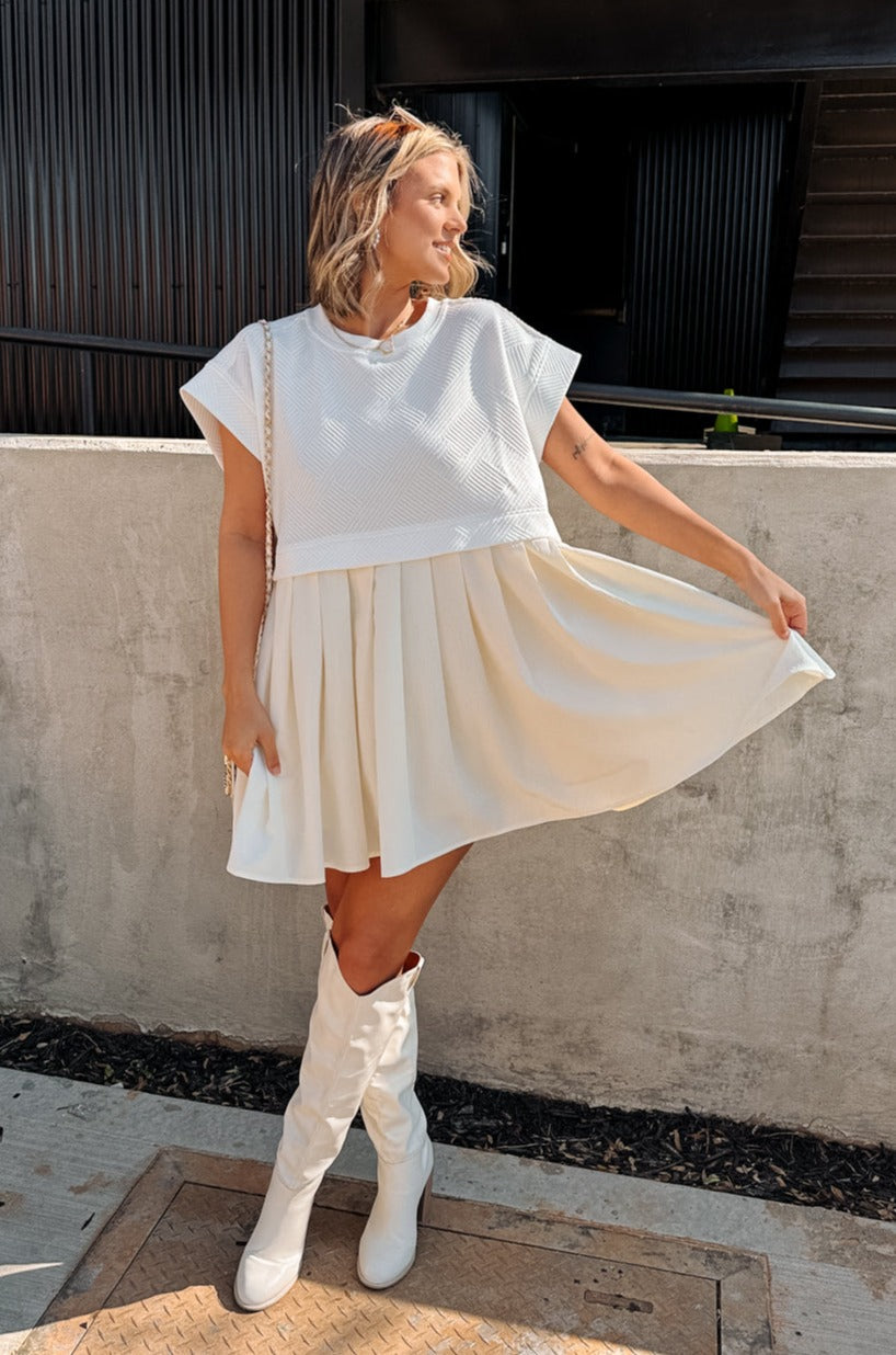 Full body front view of female model wearing the Kara Cream & White Twofer Mini Dress that has a texture white upper with short sleeves and a cream skirt with pleating. Worn with white boots.