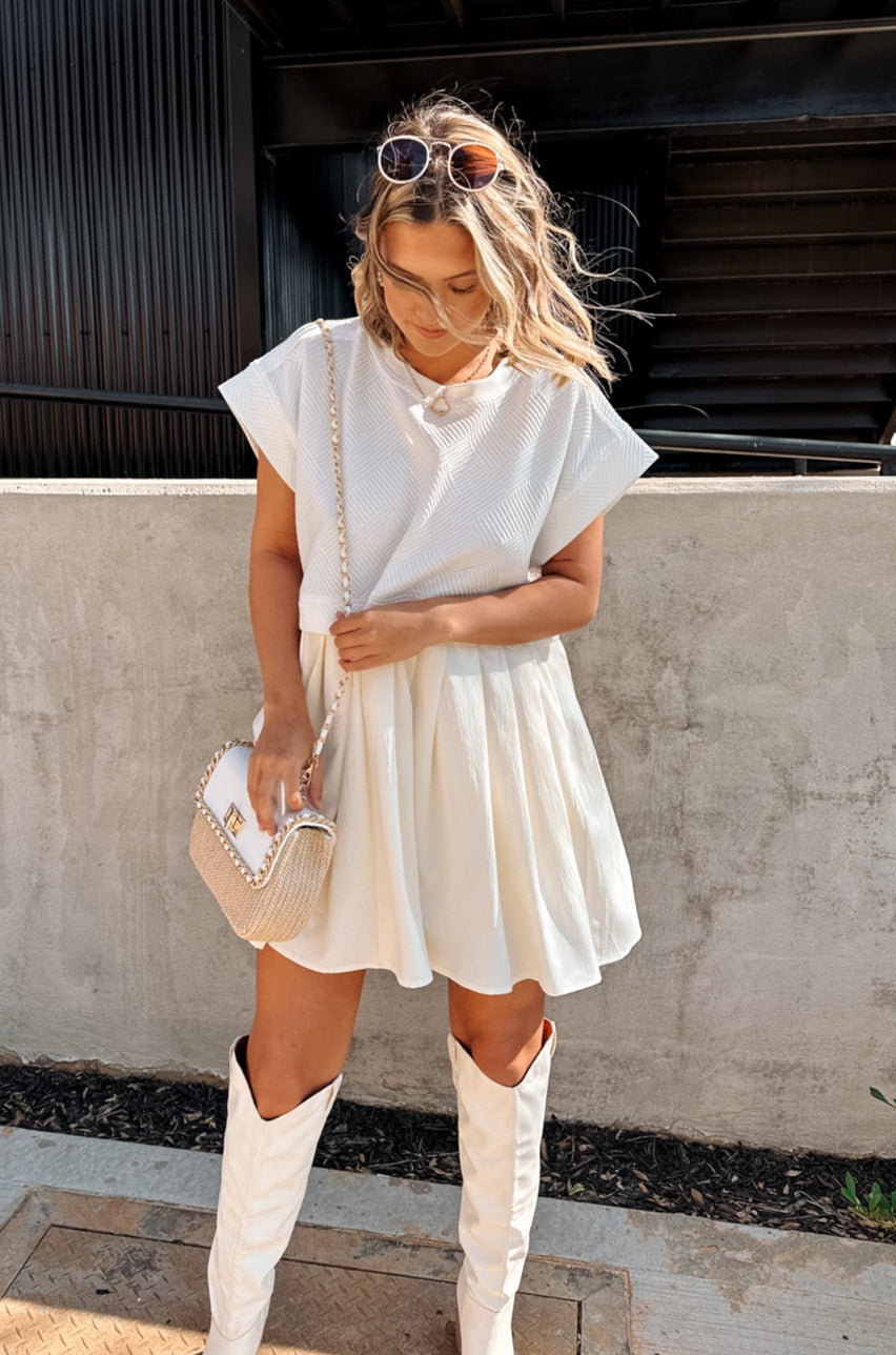 Full body front view of female model wearing the Kara Cream & White Twofer Mini Dress that has a texture white upper with short sleeves and a cream skirt with pleating. Worn with white boots.