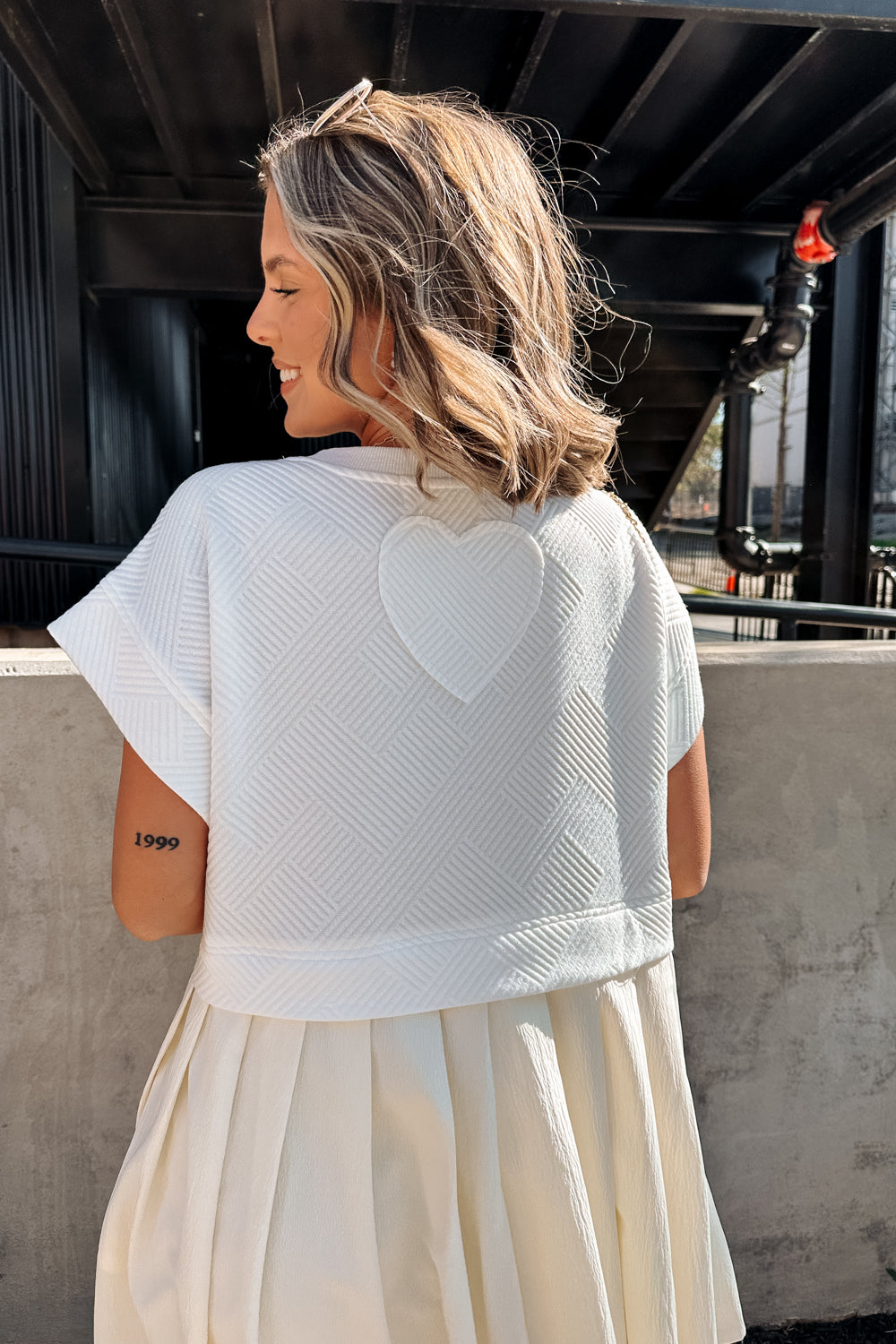 Close-up back view of female model wearing the Kara Cream & White Twofer Mini Dress that has a texture white upper with short sleeves and a cream skirt with pleating. This image shows heart patch detail on the back.