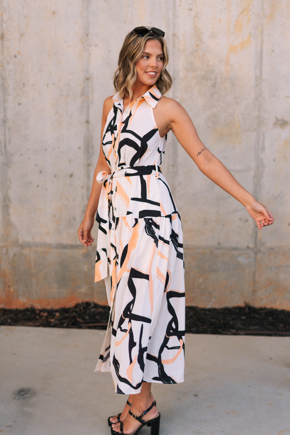 Full body side view of female model wearing the Elina Printed Button Front Midi Dress that has ivory, black, and orange geometric printing, a tie belt, a button up front, a collared sleeveless neckline, and midi length.