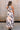 Full body back view of female model wearing the Elina Printed Button Front Midi Dress that has ivory, black, and orange geometric printing, a tie belt, a button up front, a collared sleeveless neckline, and midi length.