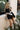 Front view of female model wearing the Livia Black Denim Dress that has black denim, tie straps, and back zipper. worn with white belt