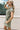 Side view of female model wearing the Abigail Olive Denim Dress that has olive denim fabric, a button up front, pockets, and collar.