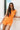 Front view of female model wearing the Raya Orange Denim Romper that has orange denim fabric, button up front, belt, and ruffle details.
