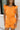 Front view of female model wearing the Raya Orange Denim Romper that has orange denim fabric, button up front, belt, and ruffle details.