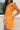 Side view of female model wearing the Raya Orange Denim Romper that has orange denim fabric, button up front, belt, and ruffle details.