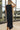 Back view of model wearing the Lillian Black Midi Dress. This is a black sleeveless midi dress with rust and cream wavy trim on sides, slit on bottom.