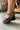 Side view of female model wearing the Cienna Wedge Sandal in Black & Natural which features Black Faux Leather, Off White Stitch Detail, Criss-Cross Straps, Adjustable Back Strap, Lug Wedge Sole and Light Brown Cushion Details