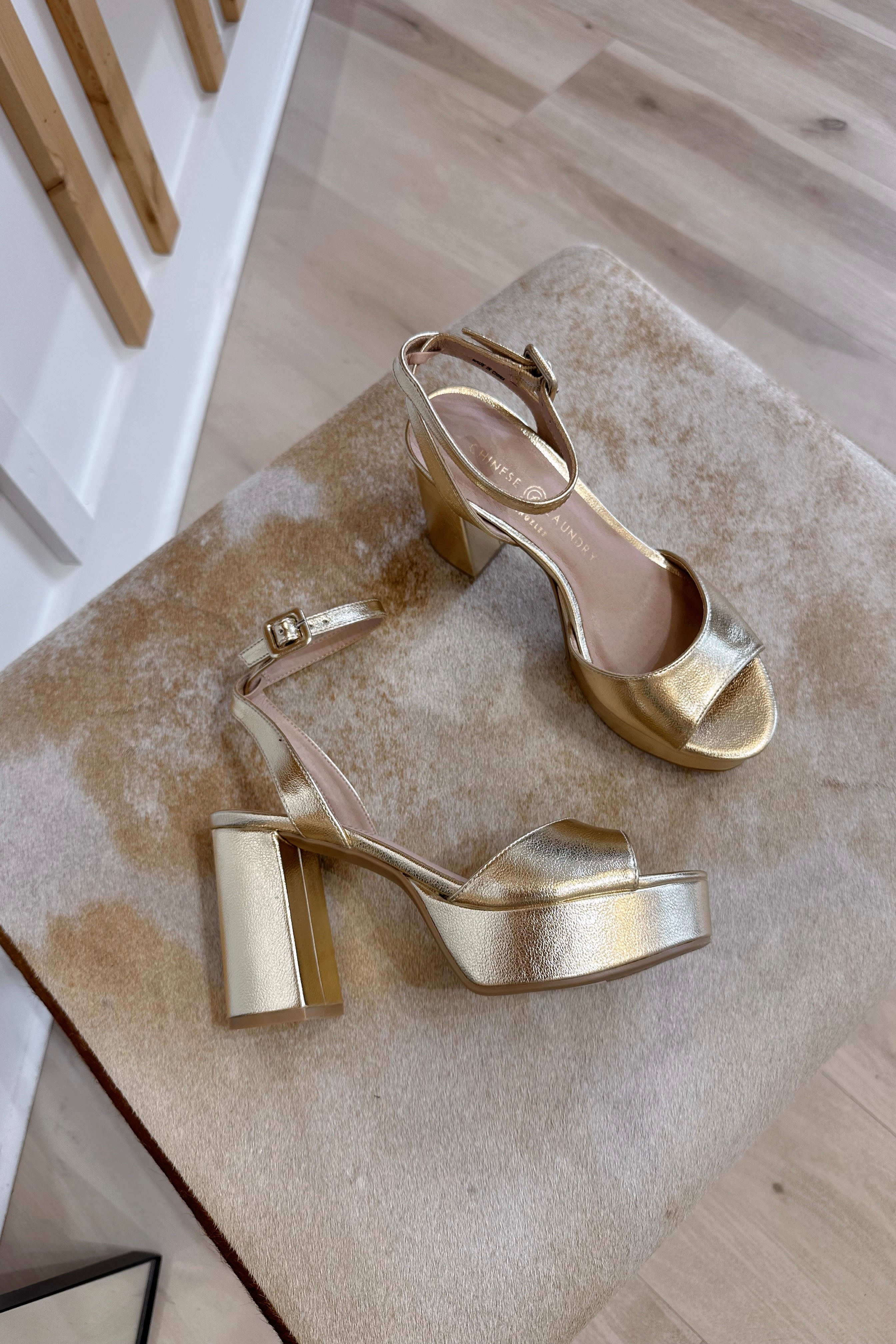 Close up detailed view of the Theresa Platform Heel in Metallic laying on a cow hide stool. The heel features gold metallic faux leather fabric, block heel, round toe, platform sole and adjustable strap.