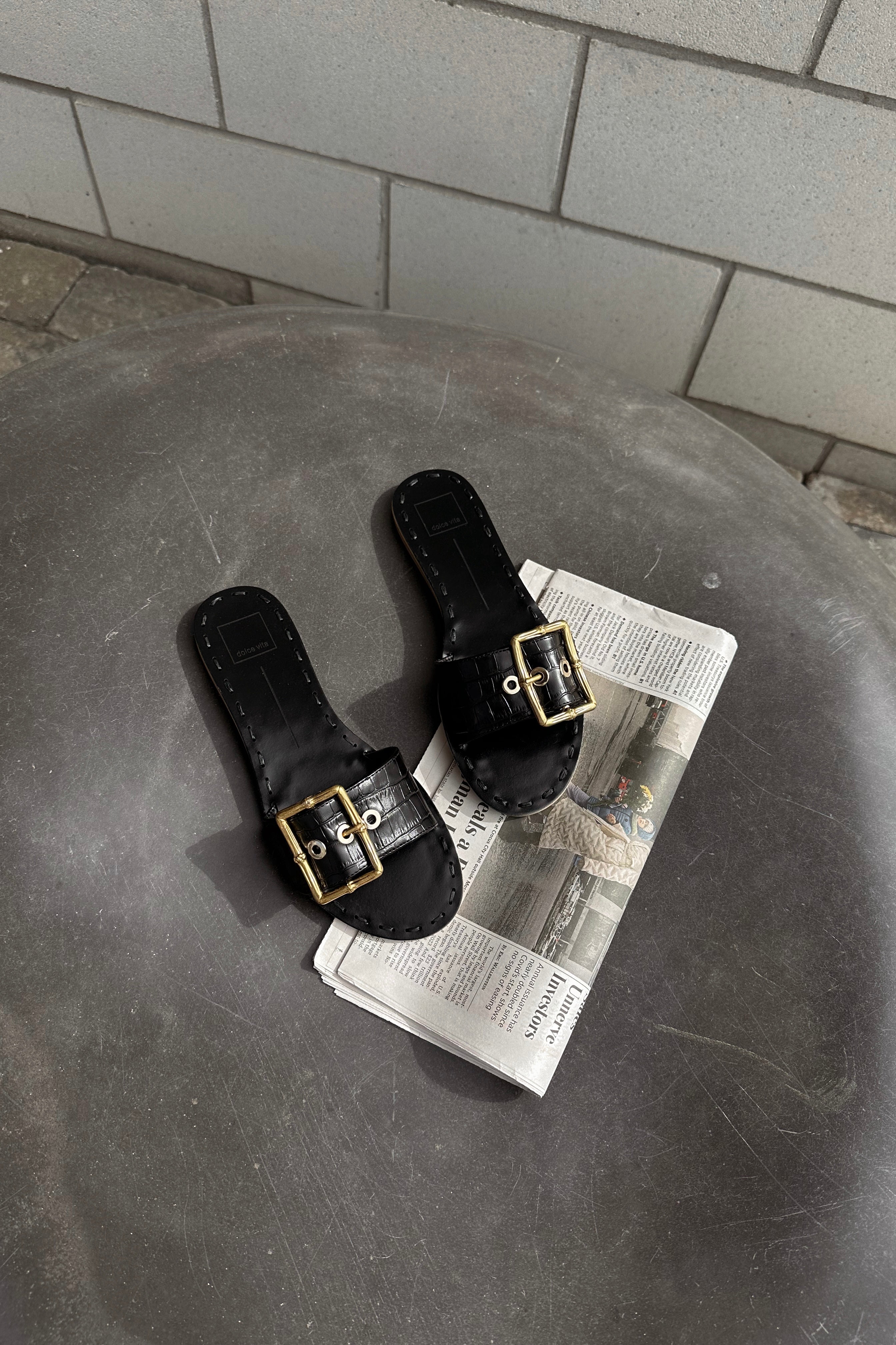 Front close up view of the Dasa Slide Sandal in Black which features Single Leather Strap, Gold Bamboo Buckle Detail and Slide-On Style. The shoe is laying over a newspaper under a black marble rock.