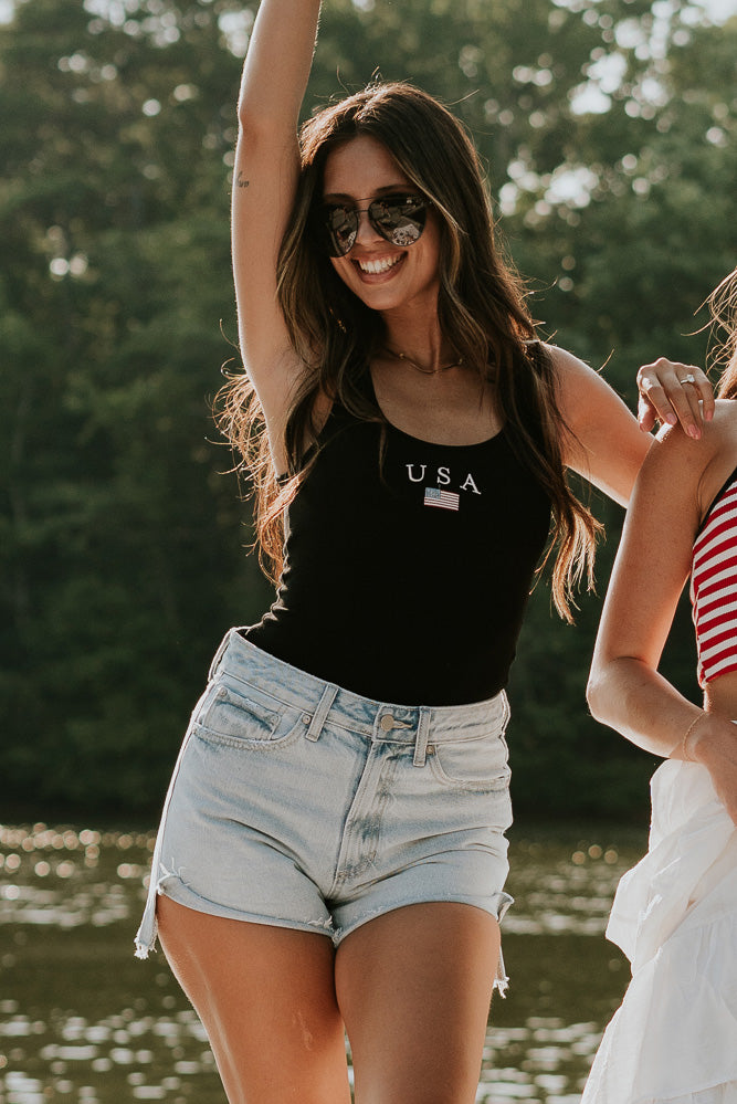Upper body front view of female model wearing the USA Black Ribbed Sleeveless Bodysuit that has black ribbed fabric, USA in white text above a flag graphic, and a scoop neck. Styled with ivory sweatshirt around shoulders and denim shorts.