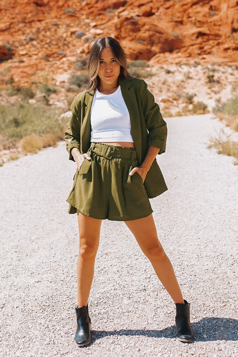 Full body frontal view of the Livin' The Dream Shorts that features an olive green colored material, a high-rise fit, an elastic waist, a belt detail, side pockets, and a flowy fit. Worn with blazer.