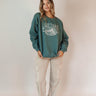 Full body front view of model wearing the Lake Tahoe Green Embroidered Sweatshirt that has heather teal green fabric, a round neckline, dropped shoulders, and long sleeves with cuffs. 