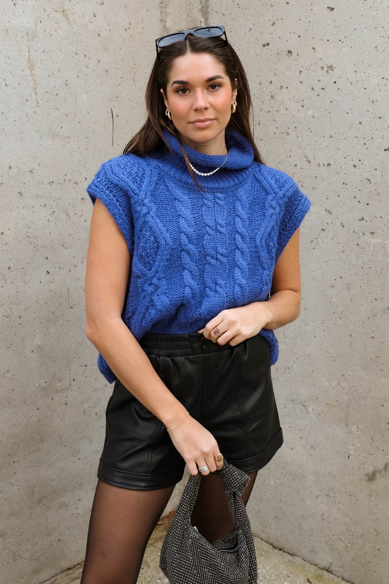 Front view of model wearing the Noelle Blue Cable Knit Sleeveless Sweater which features blue cable knit fabric, ribbed hem, high neckline and sleeveless.