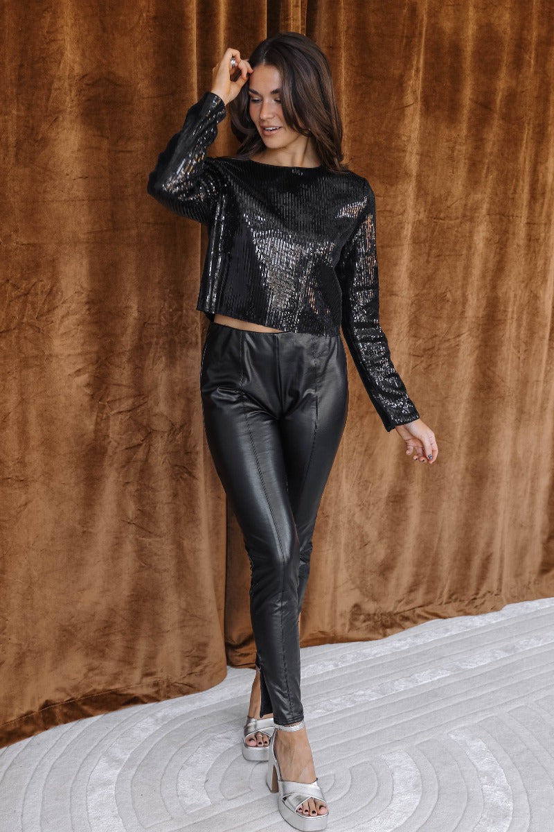 Full body front view of model wearing the Lyra Black Sequin Long Sleeve Top that has black sequin fabric, black lining, a cropped waist, a round neckline, a back zipper, and long sleeves.