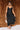 Full body front view of model wearing the Colette Black Floral Satin Midi Dress that has black satin fabric, monochrome florals, a assymetrical ruffle tier design, pleated fabric, a cowl neck, and thin straps.