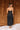 Full body back view of model wearing the Colette Black Floral Satin Midi Dress that has black satin fabric, monochrome florals, a assymetrical ruffle tier design, pleated fabric, a cowl neck, and thin straps.