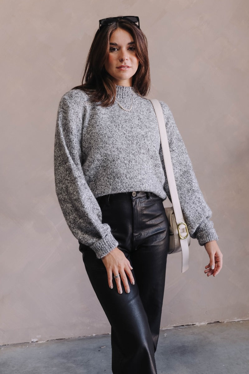 front view of model wearing the Catherine Grey Heathered Mock Neck Sweater that has charcoal grey and white textured knit fabric, a thick ribbed hem, a high neck, and long balloon sleeves.